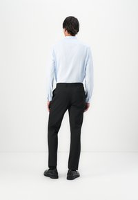Mann står med ryggen til i en lyseblå skjorte, svarte dresser, svarte sokker og svarte dressko mot en hvit bakgrunn.
