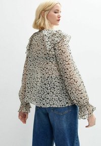Blusa nera trasparente con stampa floreale bianca, scollatura arricciata e maniche lunghe a palloncino con polsini elasticizzati; abbinata a jeans blu.