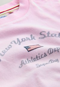 Lyserød beklædningsgenstand med teksten "New York State Athletics Dept" og en lille amerikansk flaggrafik i midten, nærbillede af stofdetalje.