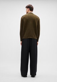 Ελιάς πράσινη πλεκτή ζακέτα με υφή, στρογγυλή λαιμόκοψη και μαύρες λεπτομέρειες στους ώμους, συνδυασμένη με χαλαρές μαύρες παντελόνες και μποτάκια.