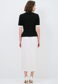 Schwarzes Peplum-Oberteil mit kurzen Ärmeln und weißen Manschetten, kombiniert mit einem weißen plissierten Midirock. Das Outfit weist eine taillierte Silhouette auf.