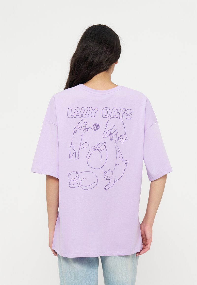 Personne aux longs cheveux foncés porte un t-shirt lavande avec l’inscription "LAZY DAYS" et des chats dessinés en contour dans différentes poses détendues au dos.