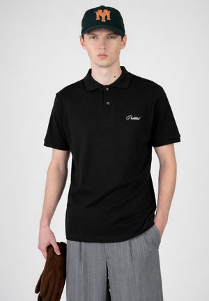 Młody mężczyzna w czarnym polo z białym napisem, szare spodnie w prążki, czarna czapka z daszkiem z pomarańczowym logo, trzymający brązowe rękawiczki w lewej ręce.