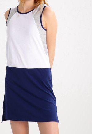 Kobieta ubrana w bezrękawnikową, casualową sukienkę z białym i szarym topem oraz granatową spódnicą, stojąca na tle jednolitego jasnego tła.