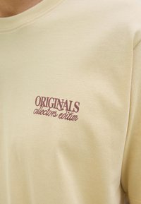 Tricou din bumbac galben deschis cu guler rotund. Prezintă text burgund: "ORIGINALS" și "ediție de colecție" pe față.