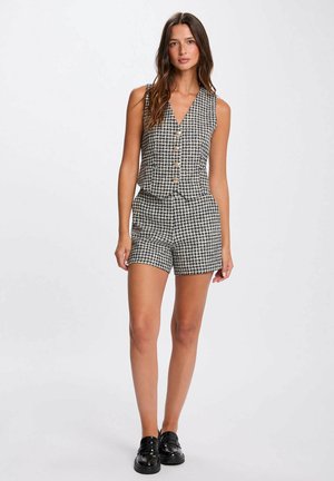 Tutina a quadri pied-de-poule in bianco e nero con scollatura profonda a V, tre bottoni e silhouette aderente, abbinata a calzature nere.