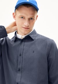 Marineblauwe blouse met knopen en kraag, met een klein geborduurd logo op de linkerkant van de borst en een gestructureerde stof. Witte onderhemd zichtbaar.