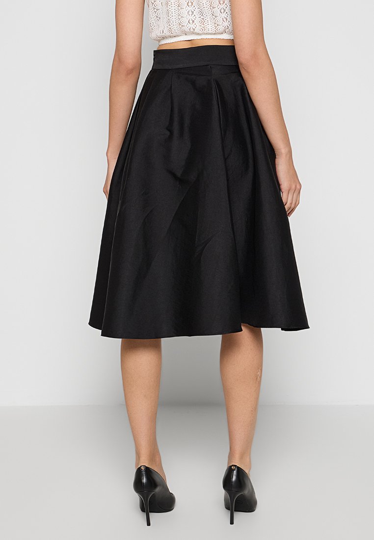 Kobieta w czarnej, do kolan, rozkloszowanej spódnicy, białej koronkowej bluzce na ramiączkach i czarnych szpilkach, stojąca tyłem do aparatu.