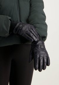 Zwarte geïsoleerde handschoenen met een gewatteerde textuur, voorzien van een logo patch, gecombineerd met een donkergroene puffer jacket en zwarte leggings.