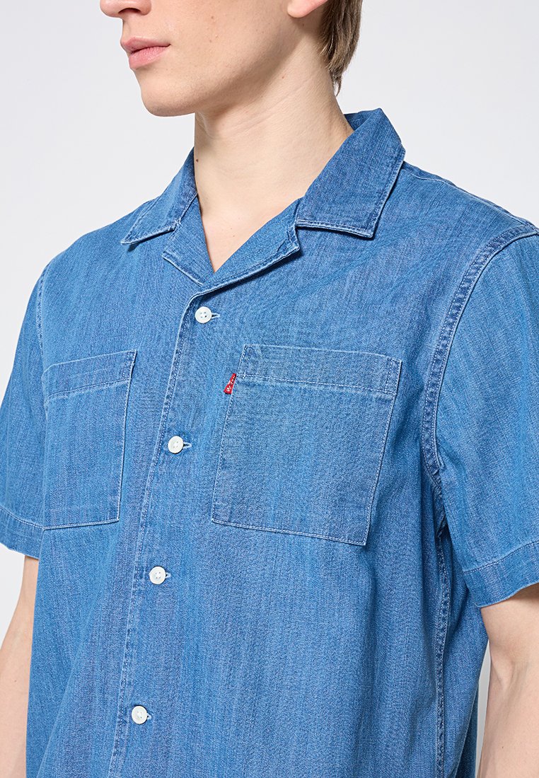 Jeune personne portant une chemise en denim à manches courtes, d'un bleu clair, avec deux poches poitrine et une petite étiquette rouge sur la poche droite.