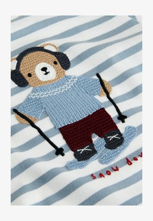 Gestreifter Stoff in Blau und Weiß zeigt einen gestickten Bären in einem blauen Pullover, roten Hosen und Skiausrüstung mit weißen Akzenten.