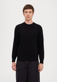 Maglione a maglia nera con motivo, scollo rotondo, maniche lunghe e polsini a costine. Indossato sopra pantaloni cargo scuri su uno sfondo neutro.