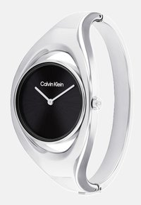 Montre Calvin Klein avec un cadran circulaire noir, un bracelet en métal argenté, un design minimaliste et des aiguillettes blanches pour les heures et les minutes. Présente le logo de la marque.