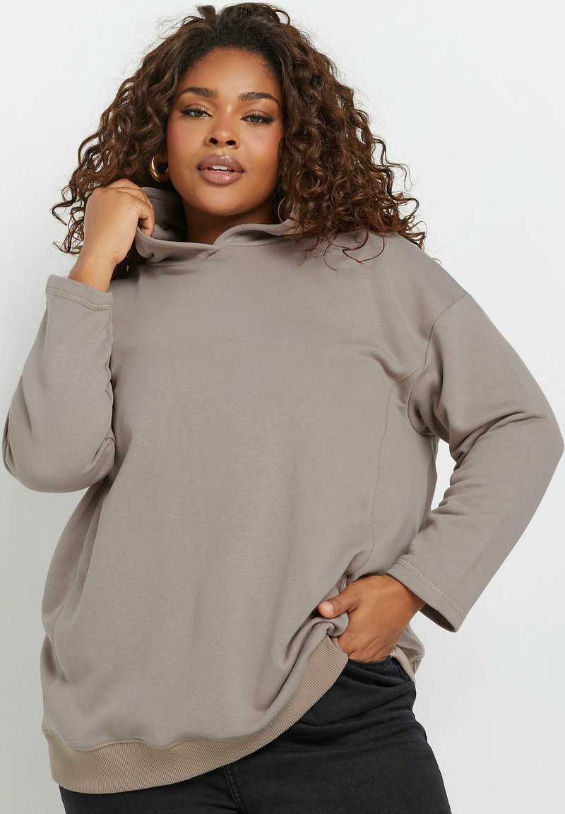 Huppari vaaleassa taupe-värissä, joka on valmistettu pehmeästä kankaasta. Siinä on pitkät hihansuut, kaareva helma ja rento istuvuus. Saumojen ympärillä on näkyvää ompelua.