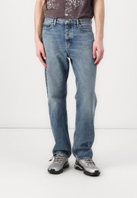 Jean en denim bleu clair avec une coupe droite, présentant un design classique à cinq poches et un léger délavage, associés à des chaussures de sport grises.