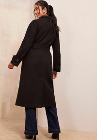 Černý dlouhý kabát s opaskem, s dlouhými rukávy a knoflíkovými manžetami. Kabát má elegantní design a je kombinován s rozšířenými džíny.