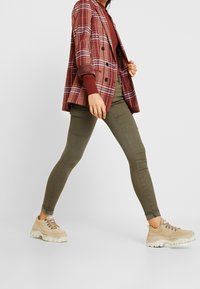 Rutigt blazer i bordeaux med vita accenter, olivgröna skinnybyxor och bruna chunky sneakers med metallisk snörhål och tjocka sulor.