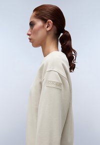 Beige sweatshirt met een zachte textuur, ronde halslijn en een prominent logo-op-patch op de linker mouw. Heeft een relaxed fit en een eenvoudig ontwerp.