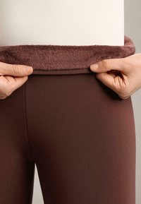 Rjavi legingi s mekim, puhastejšim bordo pasom. Roke držijo pas in prikazujejo njegovo teksturo in prileganje.