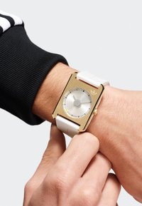 Montre rectangulaire en or avec un bracelet en caoutchouc blanc, affichant un cadran argenté, des marquages minimaux et un logo Adidas à la position de 12 heures.