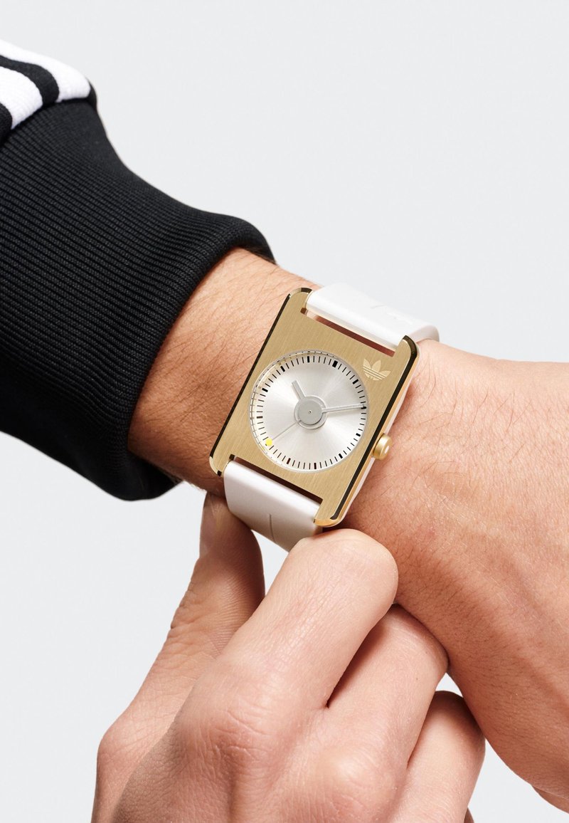 Montre rectangulaire en or avec un bracelet en caoutchouc blanc, affichant un cadran argenté, des marquages minimaux et un logo Adidas à la position de 12 heures.