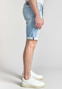 Pantalones cortos de mezclilla azul claro con dobladillos enrollados, cinco bolsillos y costura en contraste. Combinados con zapatillas blancas, mostrando un perfil lateral.