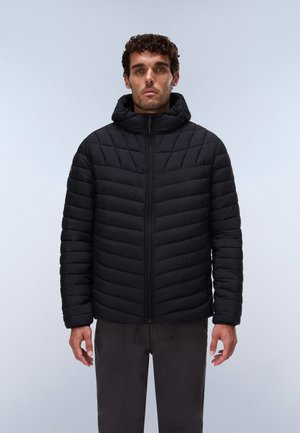 Homme portant une veste matelassée noire à capuche avec fermeture éclair devant, debout sur un fond uni gris clair.