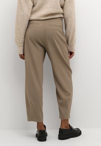 Persoon die beige op maat gemaakte enkellange broek, een beige gebreide trui en zwarte loafers met dikke zolen draagt tegen een effen witte achtergrond.