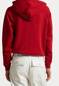 Felpa rossa con cappuccio, realizzata in tessuto morbido, con polsini a coste e una vestibilità rilassata, abbinata a pantaloni bianchi con due tasche posteriori con bottone.