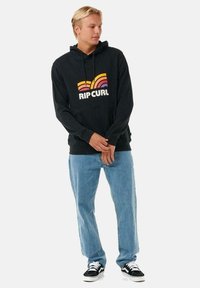 Sudadera con capucha negra con un diseño gráfico retro colorido; cuenta con un bolsillo tipo canguro, puños acanalados y un corte relajado. Combinada con unos jeans de color azul claro.