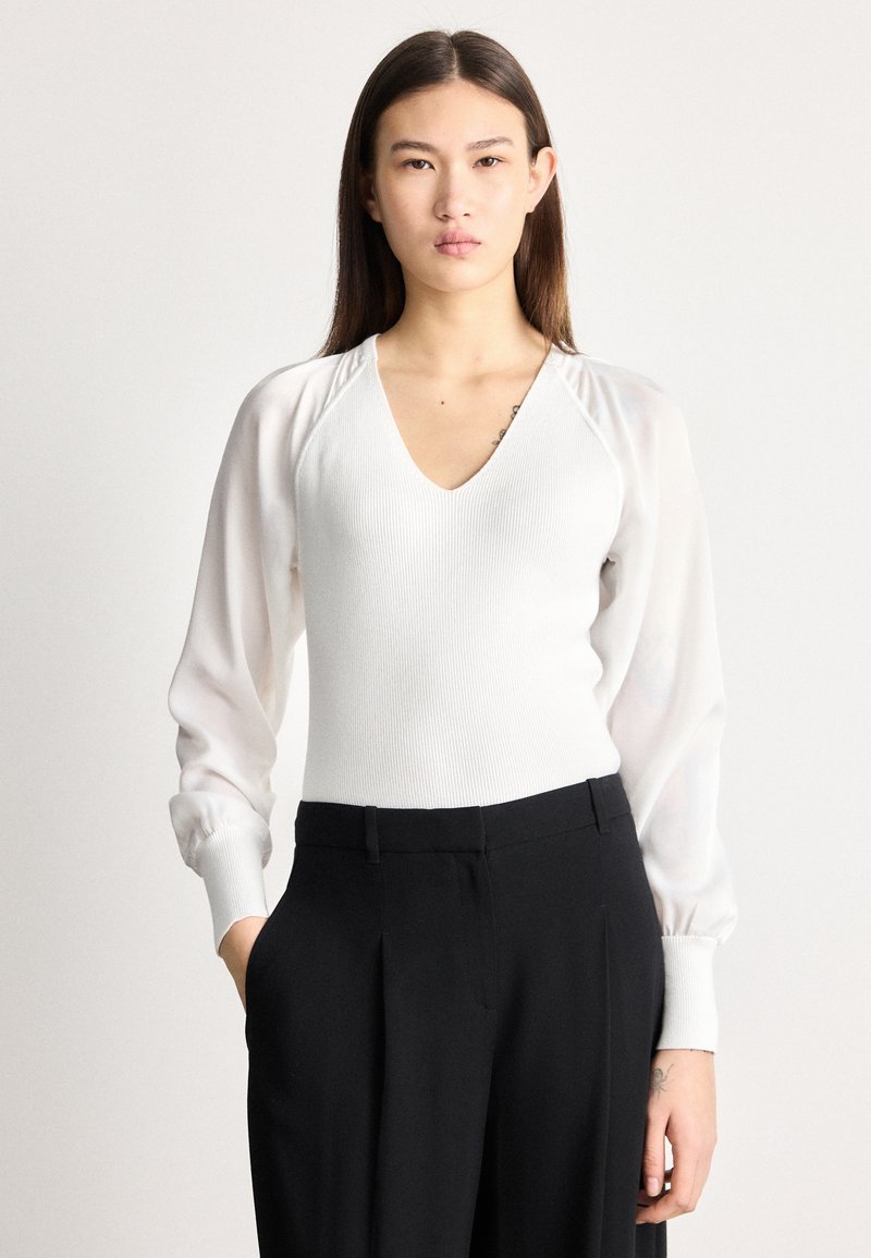 Νεαρή γυναίκα με μακριά σκούρα μαλλιά, φορούσα λευκή μπλούζα με V λαιμόκοψη και διάφανα μακριά μανίκια, συνδυασμένη με μαύρο ψηλόμεσο παντελόνι, ποζάρει με το ένα χέρι στην τσέπη.