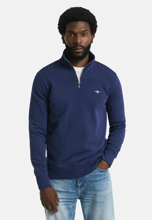 Man met een baard, gekleed in een marineblauwe kwart-rits trui over een wit shirt en lichtblauwe jeans, staand met één hand in zijn zak.