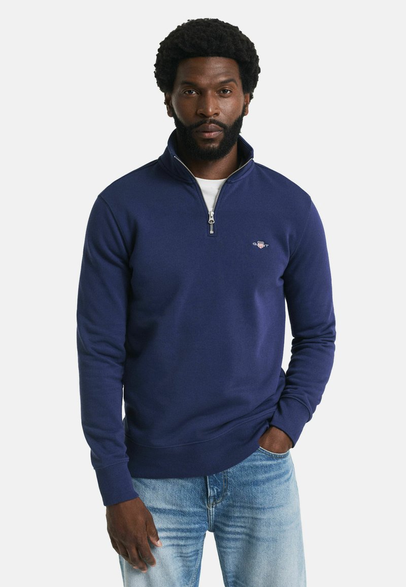 Uomo con barba che indossa un maglione a zip blu navy sopra una camicia bianca e jeans azzurri, in piedi con una mano in tasca.
