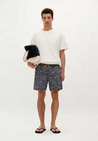 Tricou alb cu mânecă scurtă, șort de baie navy cu modele și șnur, și șlapi negri. Poartă o geantă tote albastră și crem.