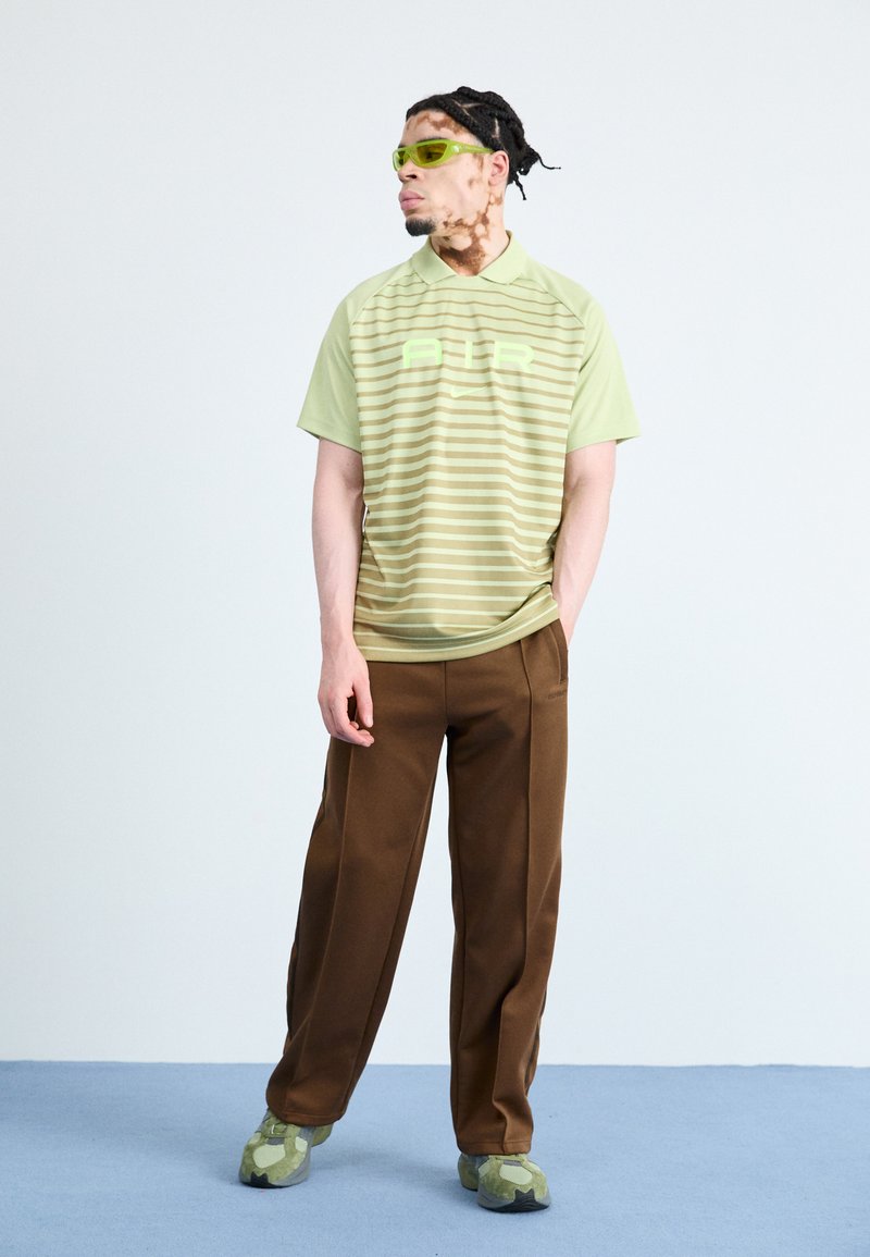 T-shirt de sport rayé vert clair avec "AIR" en vert vif, pantalon large marron, et chaussures vertes à motif camouflage. Accessoire : lunettes de soleil vertes.