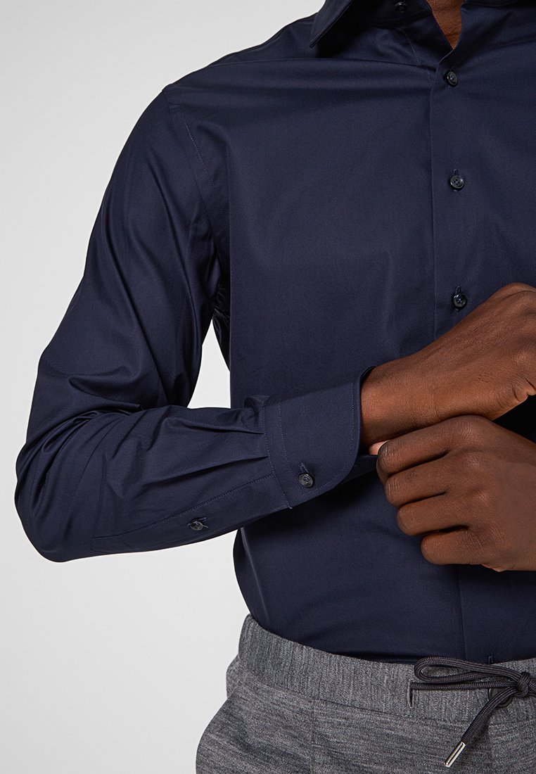 Granatowa koszula z długim rękawem, wykonana z gładkiego materiału, z kołnierzykiem zapinanym na guziki i dopasowanymi mankietami. Widać szare spodnie z elastycznym pasem.