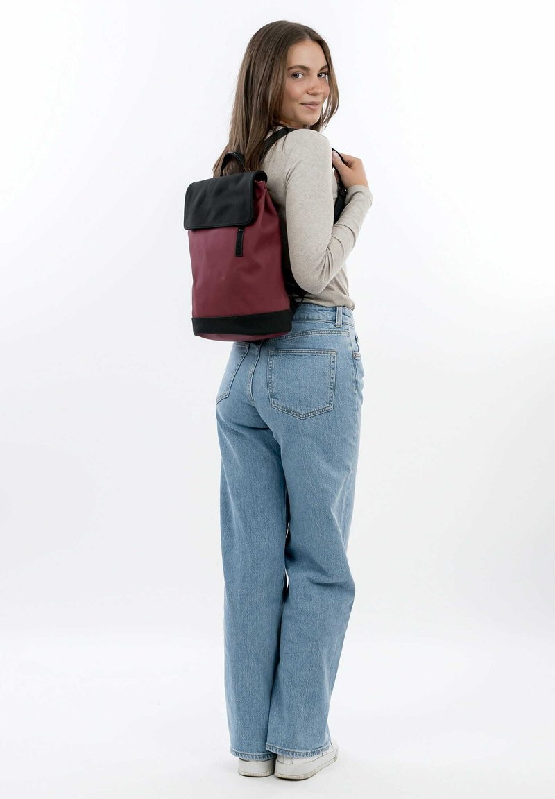 Sac à dos roulé rouge et noir avec une finition lisse, des bretelles réglables et une poche zippée à l'avant. Porté avec un jean léger bleu décontracté.