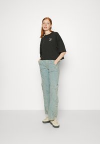 Black cropped t-shirt with Puma logo, paired with light blue cargo pants featuring multiple pockets, and beige sneakers. Simple backdrop.
