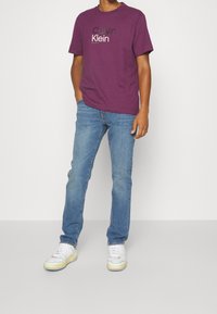 Lila bomulls T-shirt med "Calvin Klein" logotyp i vitt, kombinerad med blå denimjeans och vita sneakers med pastellaccenter.