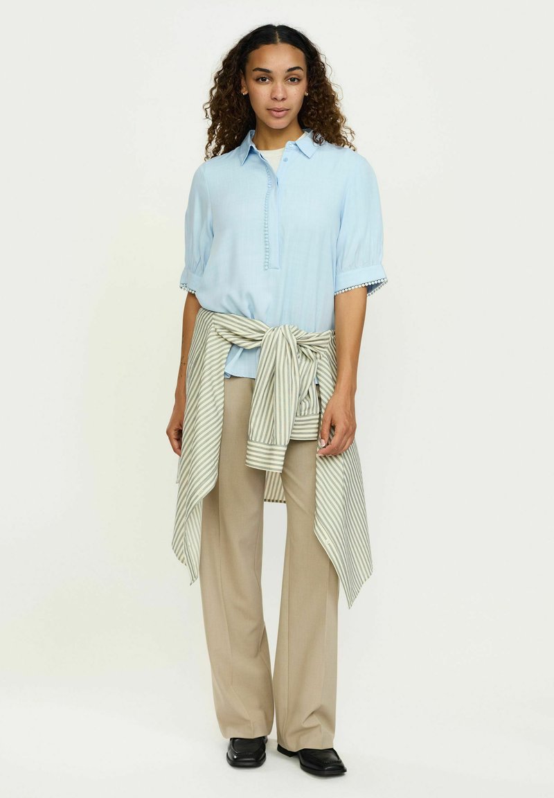 Light blue button-up shirt with short sleeves, accented with a striped gray and white wrap tied around the waist, paired with beige wide-leg trousers.
