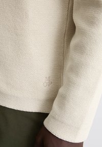 Cremfarbener Strickpullover mit strukturiertem, geripptem Design. Verfügt über eine dezente Logo-Stickerei am Saum. Die Ärmelbündchen und der Saum sind sauber verarbeitet.