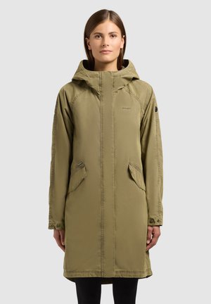 Vrouw draagt een knielange olijfgroene jas met capuchon en voorzakken, staand tegen een effen lichtgrijze achtergrond.