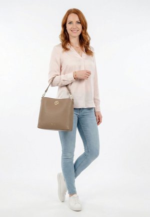 Woman with red hair wearing light pink blouse, blue jeans, and white sneakers holding a taupe handbag on her shoulder, smiling, standing on white background.