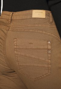 Jeans de denim castanho com um rótulo bege, apresentando um design de bolso traseiro curvado, detalhes de costura subtil e uma textura suave.
