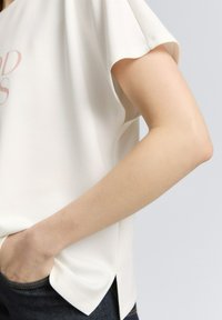 Haut à manches courtes de couleur crème avec une coupe décontractée, fentes sur les côtés et une texture lisse. Les détails incluent un motif imprimé subtil.