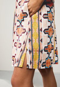 Robe multicolore avec des motifs géométriques orange, jaune et bleu sur un fond blanc ; fente latérale et détail poche.