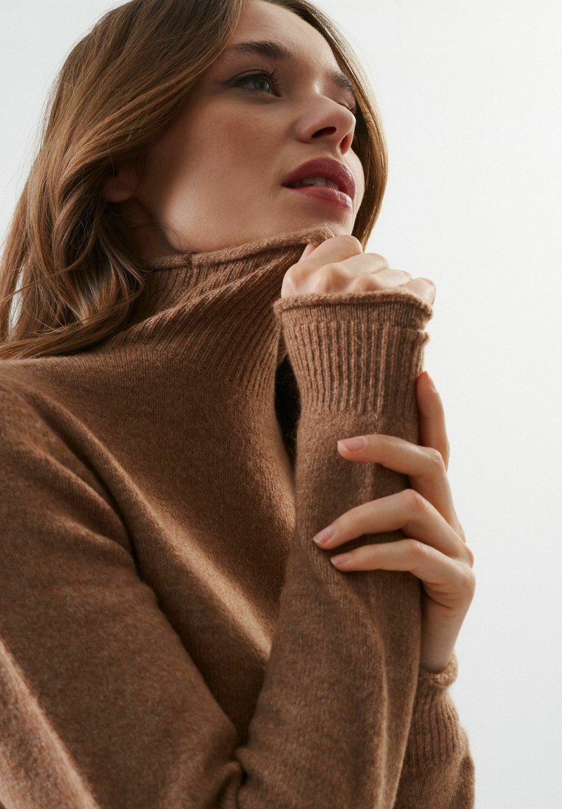 Pull en col roulé marron avec une texture côtelée et des manches ajustées, mettant en valeur un tissu doux et un col haut. Des mains tiennent délicatement le tissu.