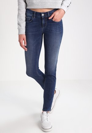 Kvinde iført mørkeblå skinny jeans, grå kort sweatshirt og hvide sneakers, stående med den ene hånd i lommen.
