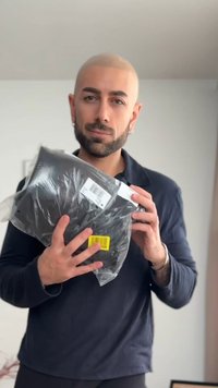 Man met korte baard en geschoren hoofd houdt een zwart, met plastic verpakt pakket met verzendlabels vast binnenshuis.