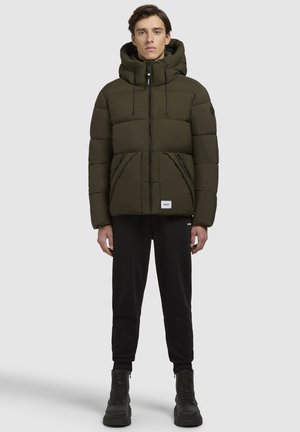 Doudoune de couleur vert foncé avec capuche, fermeture éclair sur le devant et poches latérales. Portée par-dessus un pantalon noir et des bottes noires à lacets.